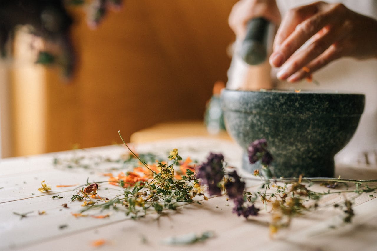 our-story Close-up of hands using mortar and pestle to grind dried flowers on wooden table.